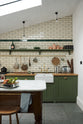 A quiet corner of a quaint and modern Wilkes green Shaker kitchen features a cooker and sink run with a rustic oak worktop, a long shaker shelf, and a Dairy table with a breakfast nook. Adorned with a long hanging rail, signature ceramics, brass cups, and a large wall of metro tiles it embodies a playful industrial and practical design.