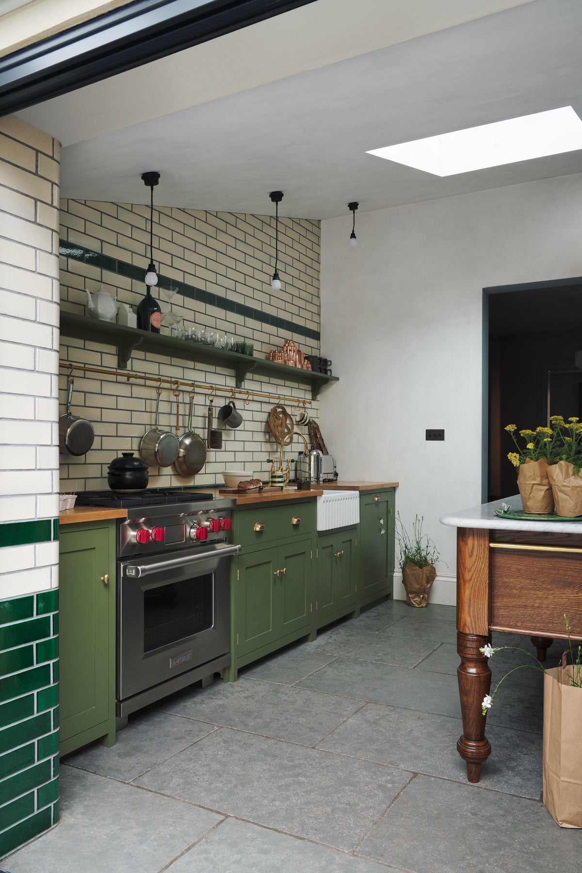 An oblique view of a quaint and modern Wilkes green Shaker kitchen features a cooker and sink run with a rustic oak worktop, a long shaker shelf, and a Dairy table with a breakfast nook. Adorned with a long hanging rail, signature ceramics, brass cups, and a large wall of metro tiles it embodies a playful industrial and practical design.
