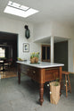 An oblique view of a quaint and modern Wilkes green Shaker kitchen features a Dairy table with a breakfast nook. Adorned with potted flowers, sky light, bum stools, and a signature glass chinese lantern inspired light it embodies a playful industrial and practical design.