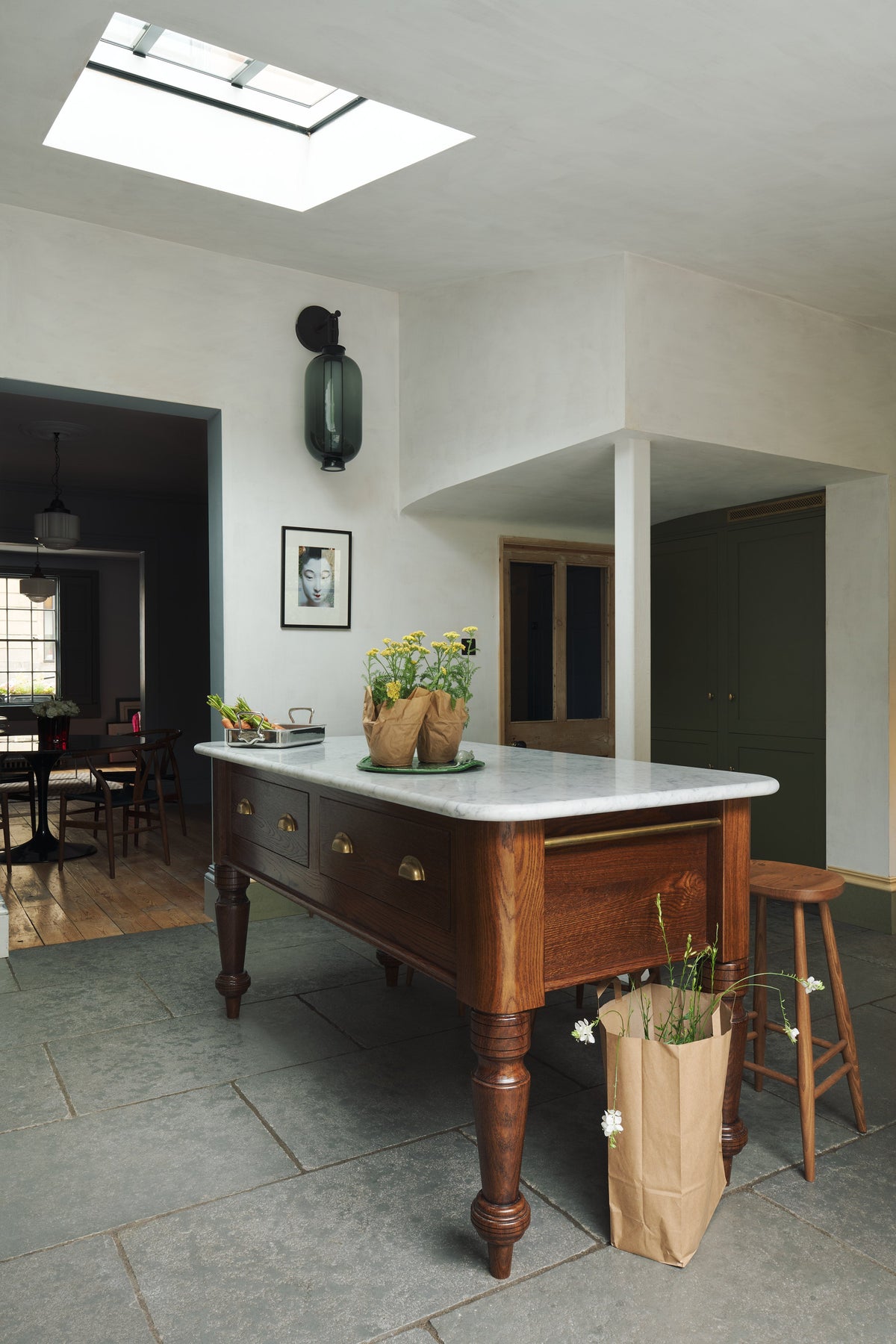 An oblique view of a quaint and modern Wilkes green Shaker kitchen features a Dairy table with a breakfast nook. Adorned with potted flowers, sky light, bum stools, and a signature glass chinese lantern inspired light it embodies a playful industrial and practical design.