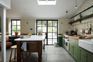 A quaint and modern Wilkes green Shaker kitchen features a cooker and sink run with a rustic oak worktop, a long shaker shelf, and a Dairy table with a breakfast nook. Adorned with a long hanging rail, sky light, glass doors, signature ceramics, brass cups, and a large wall of metro tiles it embodies a playful industrial and practical design.