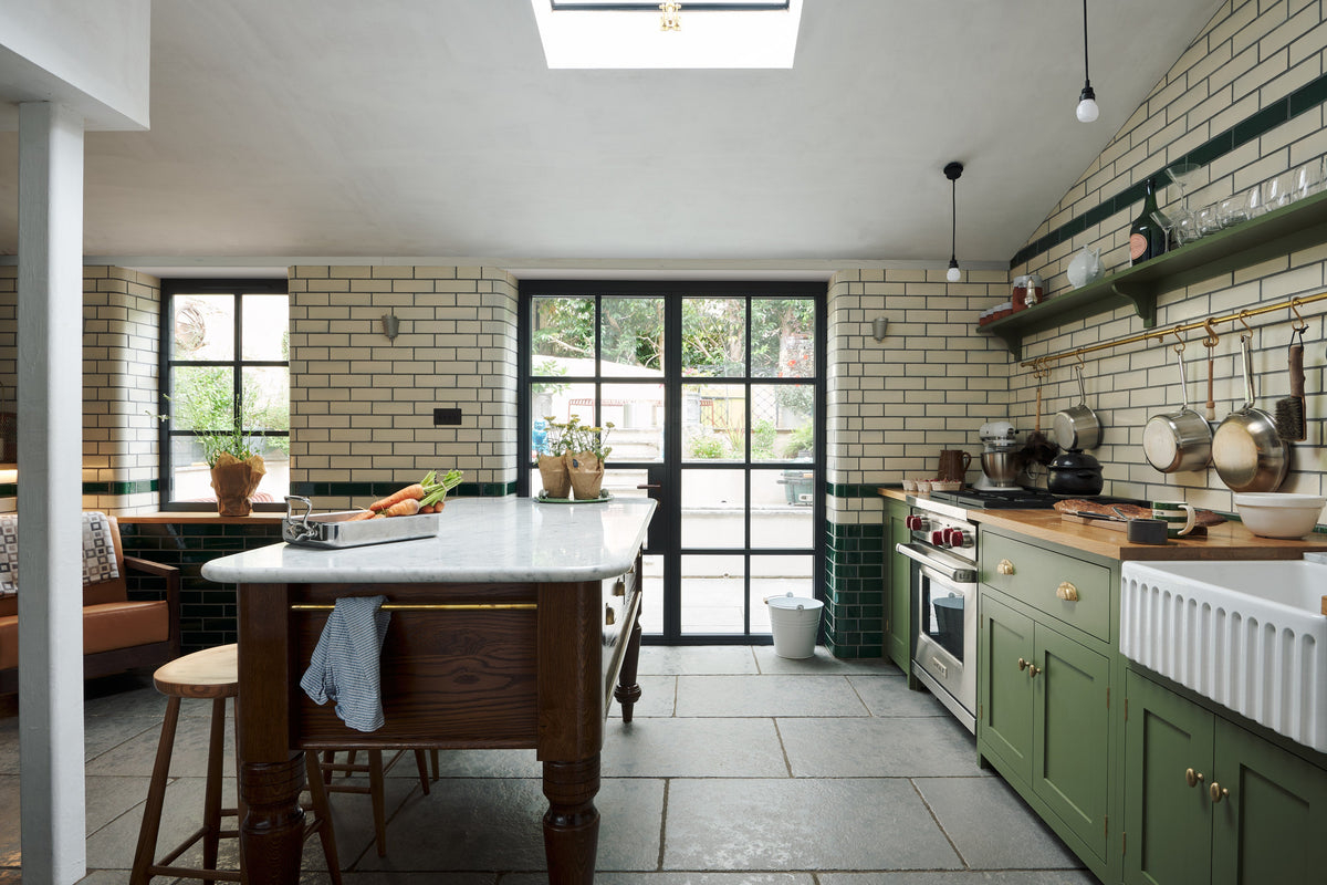 A quaint and modern Wilkes green Shaker kitchen features a cooker and sink run with a rustic oak worktop, a long shaker shelf, and a Dairy table with a breakfast nook. Adorned with a long hanging rail, sky light, glass doors, signature ceramics, brass cups, and a large wall of metro tiles it embodies a playful industrial and practical design.