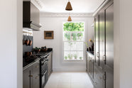 Looking through a simple archway is a murky-green galley shaker kitchen, featuring a cooker run with a black granite worktop and splashback, an aged brass hanging hook, a shaker canopy, a sink run, and pantry cupboards. Beautifully curated with two copper pendant lights, a vintage gold-framed painting, artisan ceramics, fresh produce, houseplants, and a wide open window, it creates a chic, understated, and practical galley kitchen.
