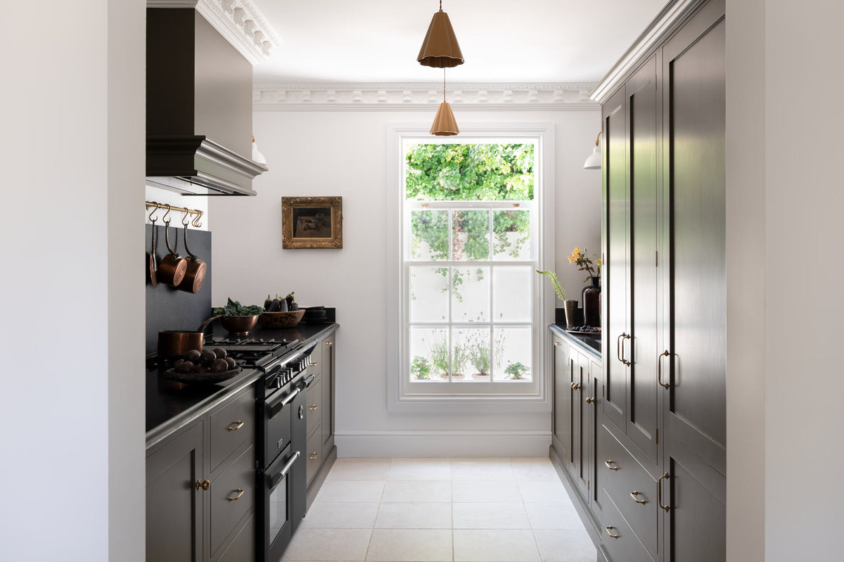 Looking through a simple archway is a murky-green galley shaker kitchen, featuring a cooker run with a black granite worktop and splashback, an aged brass hanging hook, a shaker canopy, a sink run, and pantry cupboards. Beautifully curated with two copper pendant lights, a vintage gold-framed painting, artisan ceramics, fresh produce, houseplants, and a wide open window, it creates a chic, understated, and practical galley kitchen.