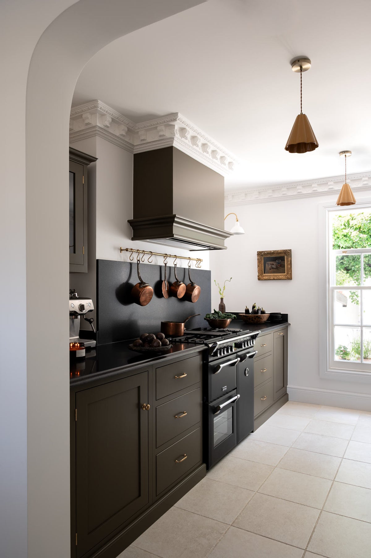 Looking through a simple archway is a murky-green galley shaker kitchen, featuring a cooker run with a black granite worktop and splashback, an aged brass hanging hook, a shaker canopy, a sink run, and pantry cupboards. Beautifully curated with two copper pendant lights, a vintage gold-framed painting, artisan ceramics, fresh produce, houseplants, and a wide open window, it creates a chic, understated, practical galley kitchen.
