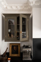 A closer look at the murky-green galley shaker kitchen, featuring a cooker run with a black granite worktop and splashback, and a glazed wall cupboard. Beautifully curated with a silver coffee machine, a vintage gold-framed painting, a vintage mortar and pestle, candle, wine glasses, and artisan ceramics, it creates a chic, understated, practical galley kitchen.