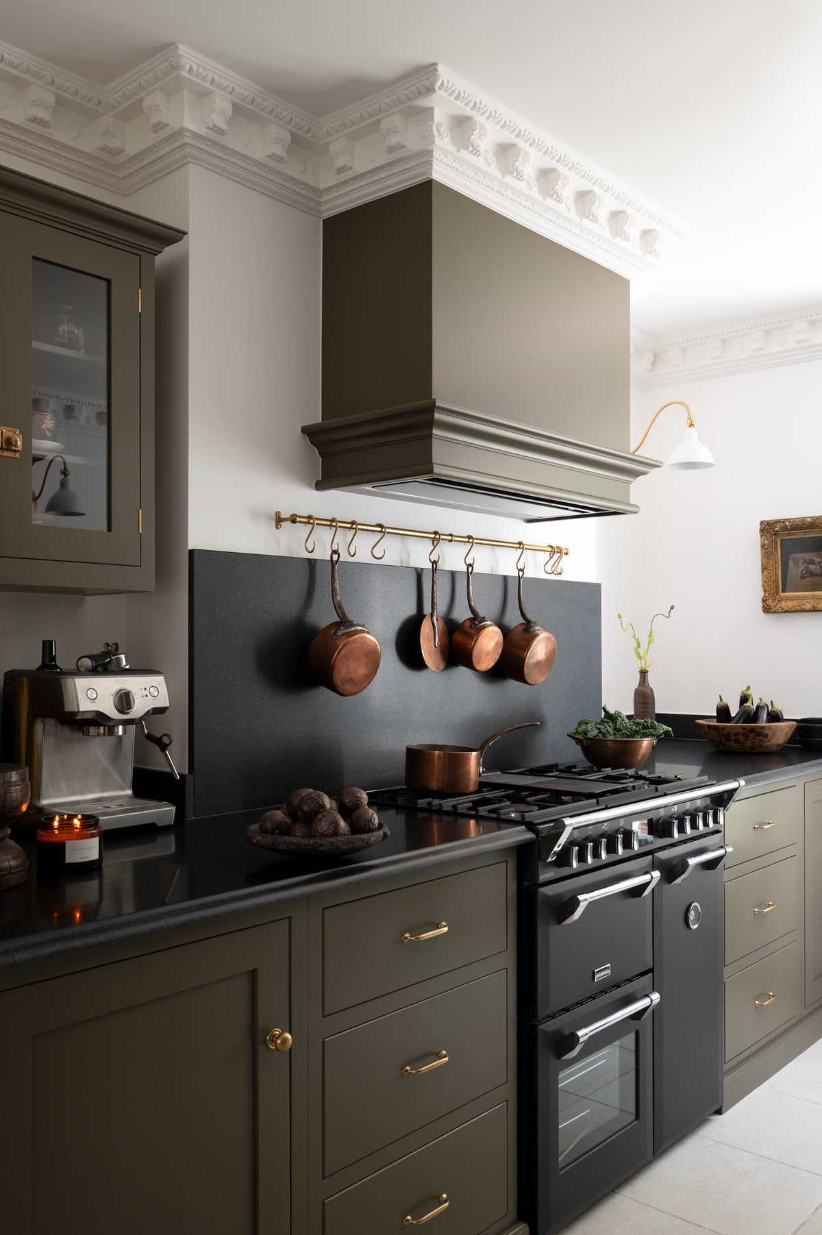 An oblique view of a murky-green galley shaker kitchen, featuring a cooker run with a black granite worktop and splashback, a shaker canopy, a glazed wall cupboard, and an aged brass hanging rail. Beautifully curated with a vintage gold-framed painting, artisan ceramics, fresh produce, houseplants, and copper pans, it creates a chic, understated, practical galley kitchen.