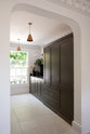 An oblique view through a simple archway is a murky-green galley shaker kitchen, featuring a sink run with a black granite worktop and splashback, a pantry cupboard, and a shaker shelf with pegs. Beautifully curated with two copper pendant lights, artisan ceramics, fresh houseplants, and a wide open window, it creates a chic, understated, and practical galley kitchen.