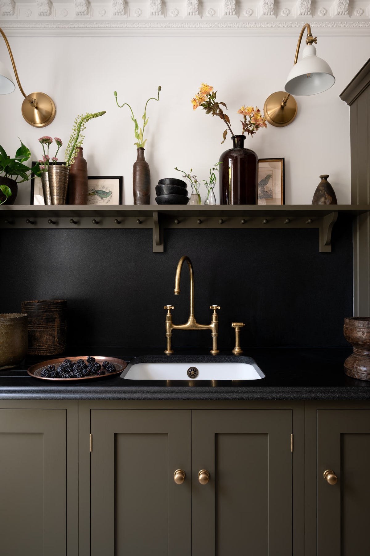 A closer look at the murky-green galley shaker kitchen, featuring a sink run with a black granite worktop and splashback, an undermounted sink, an aged brass ionian tap, and a shaker shelf with pegs. Beautifully curated with two brass wall lights, artisan ceramics, fresh houseplants, framed vintage illustrations, and a brass plate of blackberries, it creates a chic, understated, and practical galley kitchen.