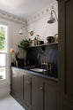 An oblique view of the murky-green galley shaker kitchen, featuring a sink run with a black granite worktop and splashback, an undermounted sink, an aged brass ionian tap, and a shaker shelf with pegs. Beautifully curated with two brass wall lights, artisan ceramics, fresh houseplants, framed vintage illustrations, and a brass plate of blackberries, it creates a chic, understated, and practical galley kitchen.