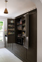 An oblique view through a simple archway is a murky-green galley shaker kitchen, featuring a sink run with a black granite worktop and splashback, a pantry cupboard, and a shaker shelf with pegs. Beautifully curated with two copper pendant lights, artisan ceramics, fresh houseplants, and a wide open window, it creates a chic, understated, and practical galley kitchen.