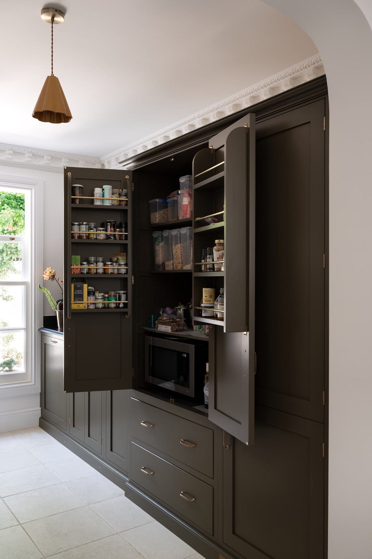 An oblique view through a simple archway is a murky-green galley shaker kitchen, featuring a sink run with a black granite worktop and splashback, a pantry cupboard, and a shaker shelf with pegs. Beautifully curated with two copper pendant lights, artisan ceramics, fresh houseplants, and a wide open window, it creates a chic, understated, and practical galley kitchen.