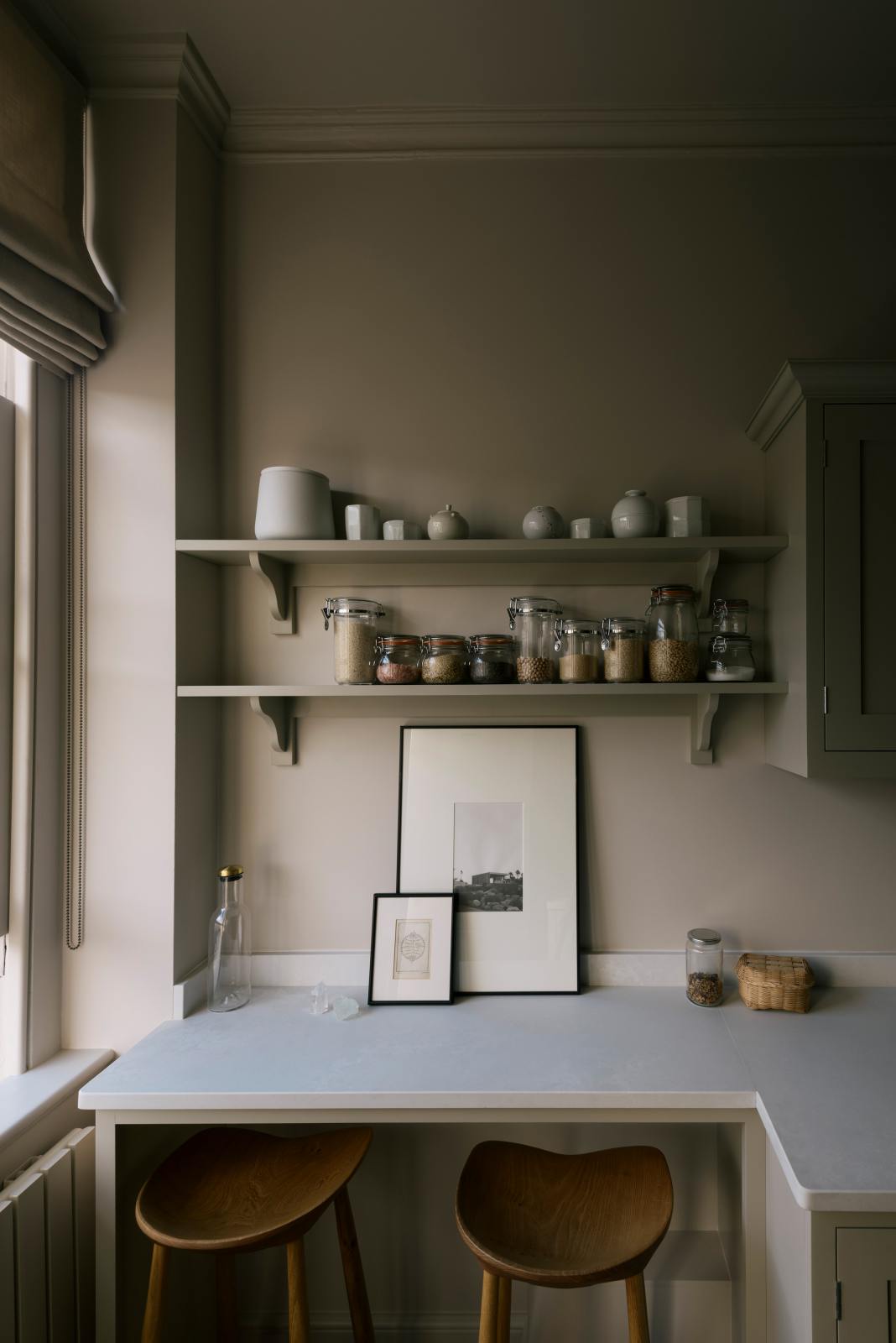 A stylish, mushroom-coloured Shaker galley kitchen features a cooker and sink run with Silestone worktop, wall cupboards and a cosy breakfast nook. Beautifully decorated with simple, everyday kitchen items such as dried ingredients, muted ceramics, framed illustrations, deVOL Bum Chairs, a healing crystal, and an amazing view of Bath’s famous Georgian apartments, this calming and functional kitchen space blends character with peace.