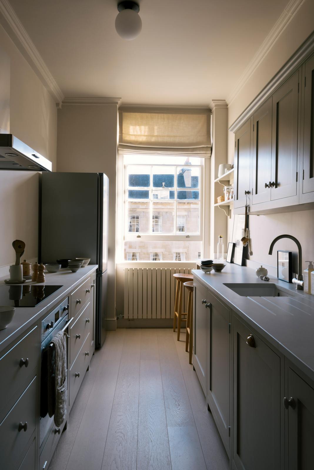 Nestled between a cooker and sink run with Silestone worktop, wall cupboards and a cosy breakfast nook of a stylish, mushroom-coloured Shaker galley kitchen. Beautifully decorated with simple, everyday kitchen items such as dried ingredients, muted ceramics, framed illustrations, deVOL Bum Chairs, and an amazing view of Bath’s famous Georgian apartments, this calming and functional kitchen space blends character with peace.