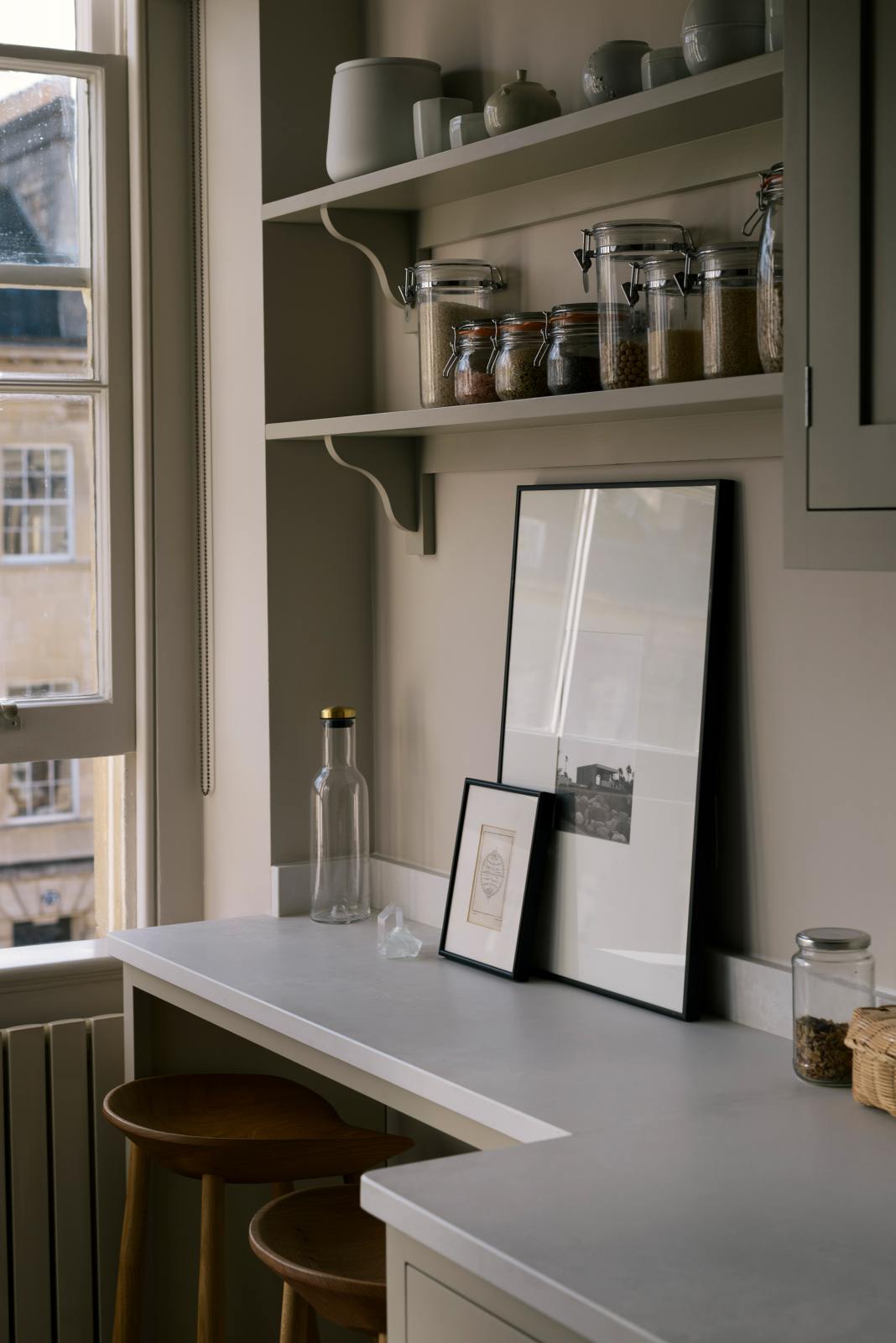 An angled view of a stylish, mushroom-coloured Shaker galley kitchen features a cooker and sink run with Silestone worktop, wall cupboards and a cosy breakfast nook. Beautifully decorated with simple, everyday kitchen items such as dried ingredients, muted ceramics, framed illustrations, deVOL Bum Chairs, a healing crystal, and an amazing view of Bath’s famous Georgian apartments, this calming and functional kitchen space blends character with peace.