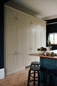 Nestled between a simple and elegant mushroom-brown Shaker kitchen, it features a large kitchen island with a single Belfast sink, a small section of copper worktop, and a bank of fitted cupboards housing a fridge-freezer and storage pantries. Adorned with artisan ceramics, gem lettuce, bright red rhubarb, luscious houseplants, and a deVOL Heirloom gaselier, it creates a loving and functional space.