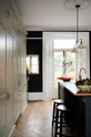 Nestled between a simple and elegant mushroom-brown Shaker kitchen, it features a large kitchen island with a single Belfast sink, a small section of copper worktop, and a bank of fitted cupboards housing a fridge-freezer and storage pantries. Adorned with artisan ceramics, gem lettuce, bright red rhubarb, luscious houseplants, and a deVOL Heirloom gaselier, it creates a loving and functional space.