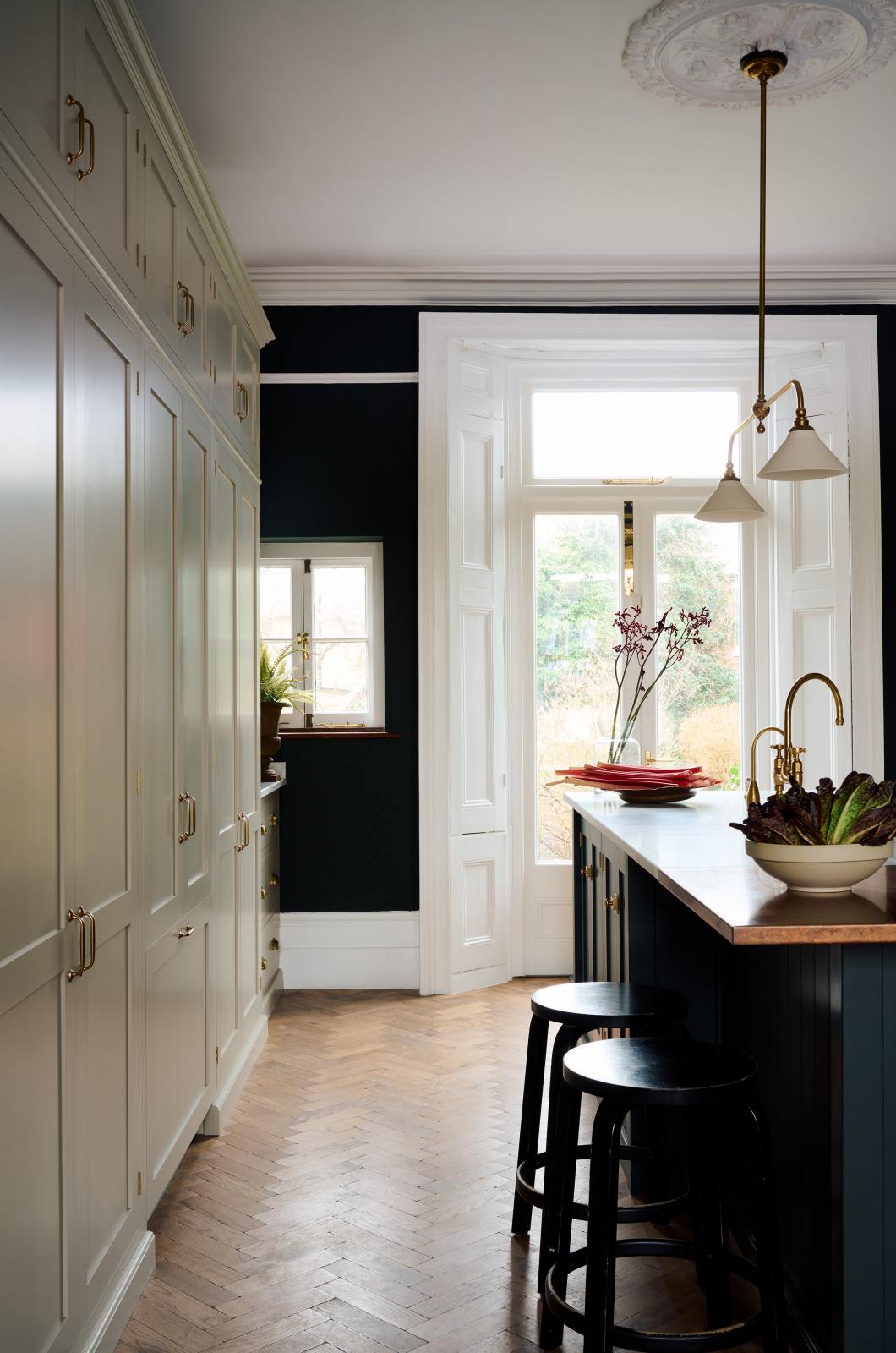 Nestled between a simple and elegant mushroom-brown Shaker kitchen, it features a large kitchen island with a single Belfast sink, a small section of copper worktop, and a bank of fitted cupboards housing a fridge-freezer and storage pantries. Adorned with artisan ceramics, gem lettuce, bright red rhubarb, luscious houseplants, and a deVOL Heirloom gaselier, it creates a loving and functional space.