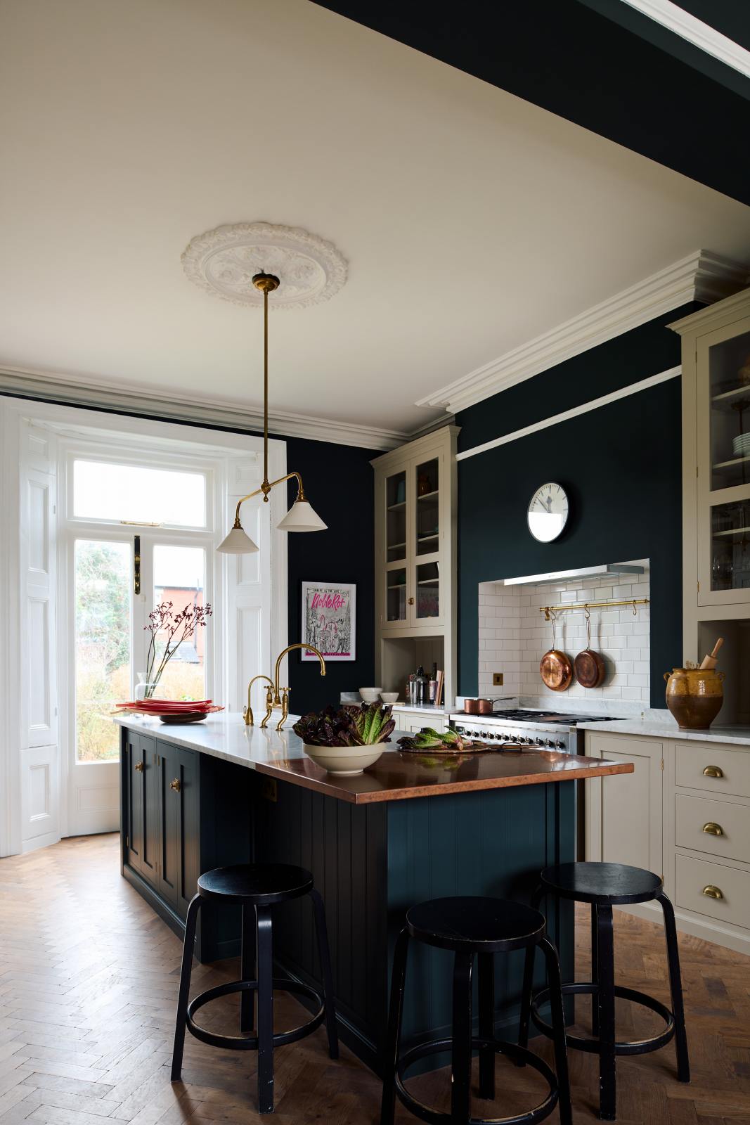 An oblique view of a simple and elegant mushroom-brown Shaker kitchen features a cooker run with freestanding cupboards, a Carrara marble worktop, a large kitchen island with a single Belfast sink, and a small section of copper worktop. Adorned with artisan ceramics, a playful framed illustration, gem lettuce, bright red rhubarb, elegant florals in a vase, and a deVOL Heirloom gaselier, the space feels loving and functional.