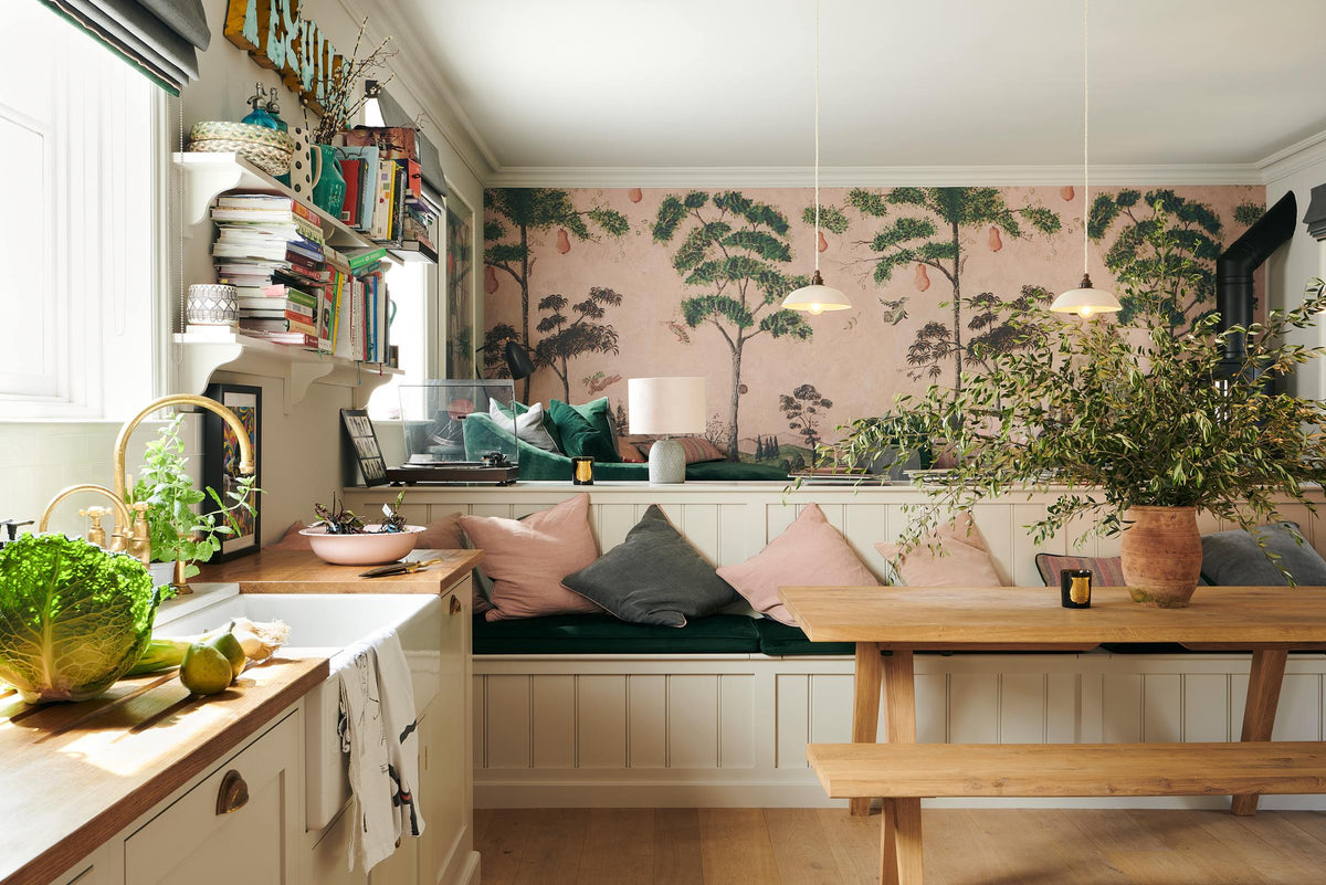 A vibrant Shaker kitchen features an L-shaped sink and cooker run with a solid oak worktop, a single Belfast sink, an aged brass Ionian tap, a Shaker shelf, and a bench. Lovingly decorated with pink rainforest wallpaper, colourful cushions, overstocked books, a record player, chattered skullcap porcelain lights, and a park bench dining table, it creates a characterful and inviting space.