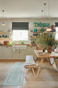 A vibrant Shaker kitchen features an L-shaped sink and cooker run with a solid oak worktop, a single Belfast sink, an aged brass Ionian tap, a Shaker shelf, and a bench. Lovingly decorated with a vintage “Tequila” sign, colourful cushions, overstocked books, a wild fauna arrangement, chattered skullcap porcelain lights, and a park bench dining table, it creates a characterful and inviting space.