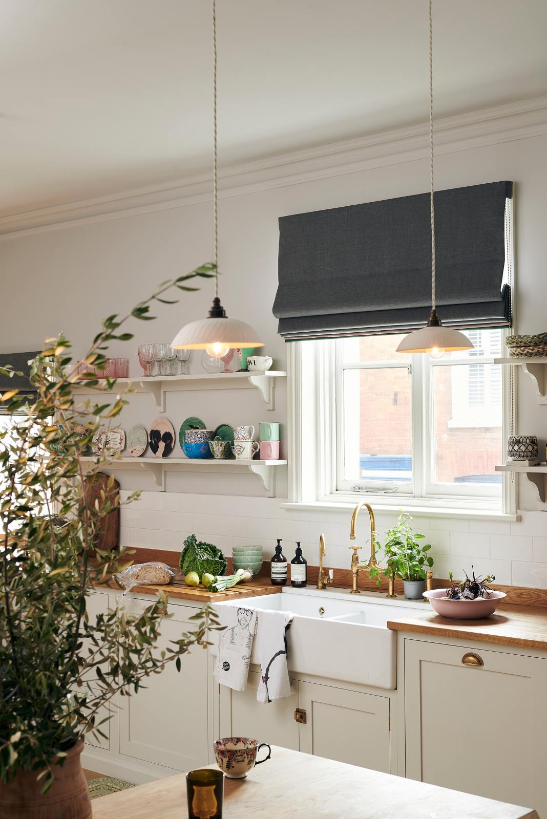 An oblique view of a vibrant Shaker kitchen features an L-shaped sink and cooker run with a solid oak worktop, a single Belfast sink, an aged brass Ionian tap, and Shaker shelves. Lovingly decorated with beautiful ceramics and drinking glasses, luscious green vegetables, and chattered skullcap porcelain lights, it creates a characterful and inviting space.