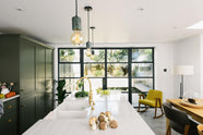 A bright and sophisticated Bakehouse Green shaker kitchen, styled with Scandinavian furniture and a soothing green palette. The kitchen features a cooker run, a central island with a double Belfast sink and Silestone worktop, and full-height cupboards. Natural light floods the kitchen and dining room through the glass French doors.