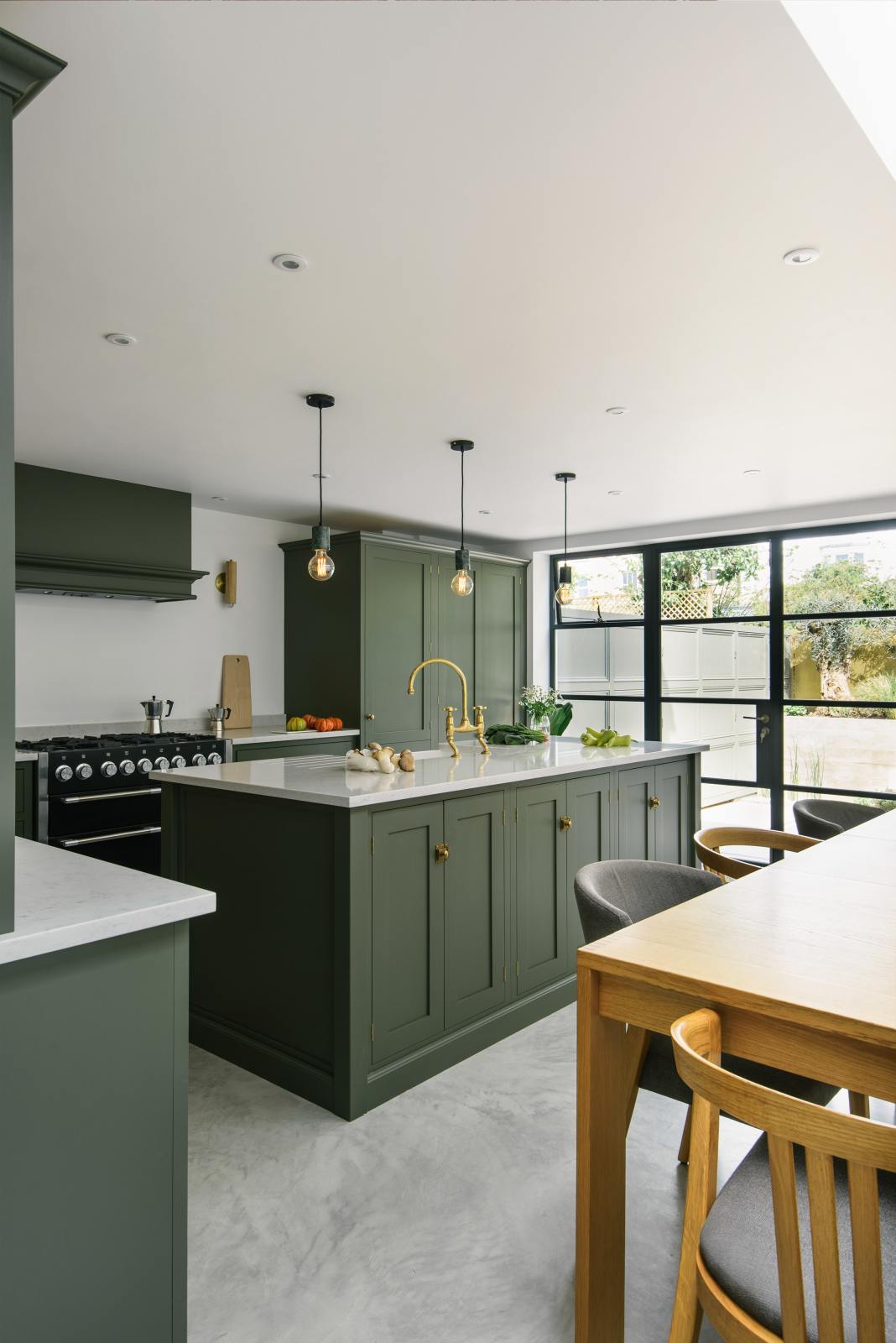 An angled view of a bright and sophisticated Bakehouse Green shaker kitchen, styled with Scandinavian furniture and a soothing green palette. The kitchen features a cooker run, a central island with a double Belfast sink and Silestone worktop, and full-height cupboards. Natural light floods the kitchen and dining room through the glass French doors.