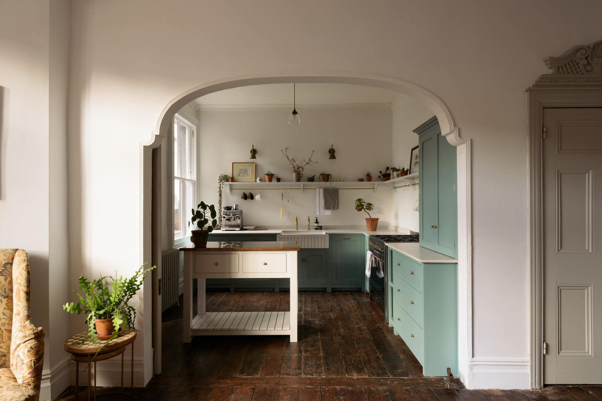 A beautifully detailed alcove features a Trinity Blue shaker kitchen with an L-shaped cooker and sink run. The kitchen includes a countertop cupboard topped with Carrara marble and a freestanding prep table in linen white with a copper worktop. The setting is in a well-preserved house with its original floorboards.