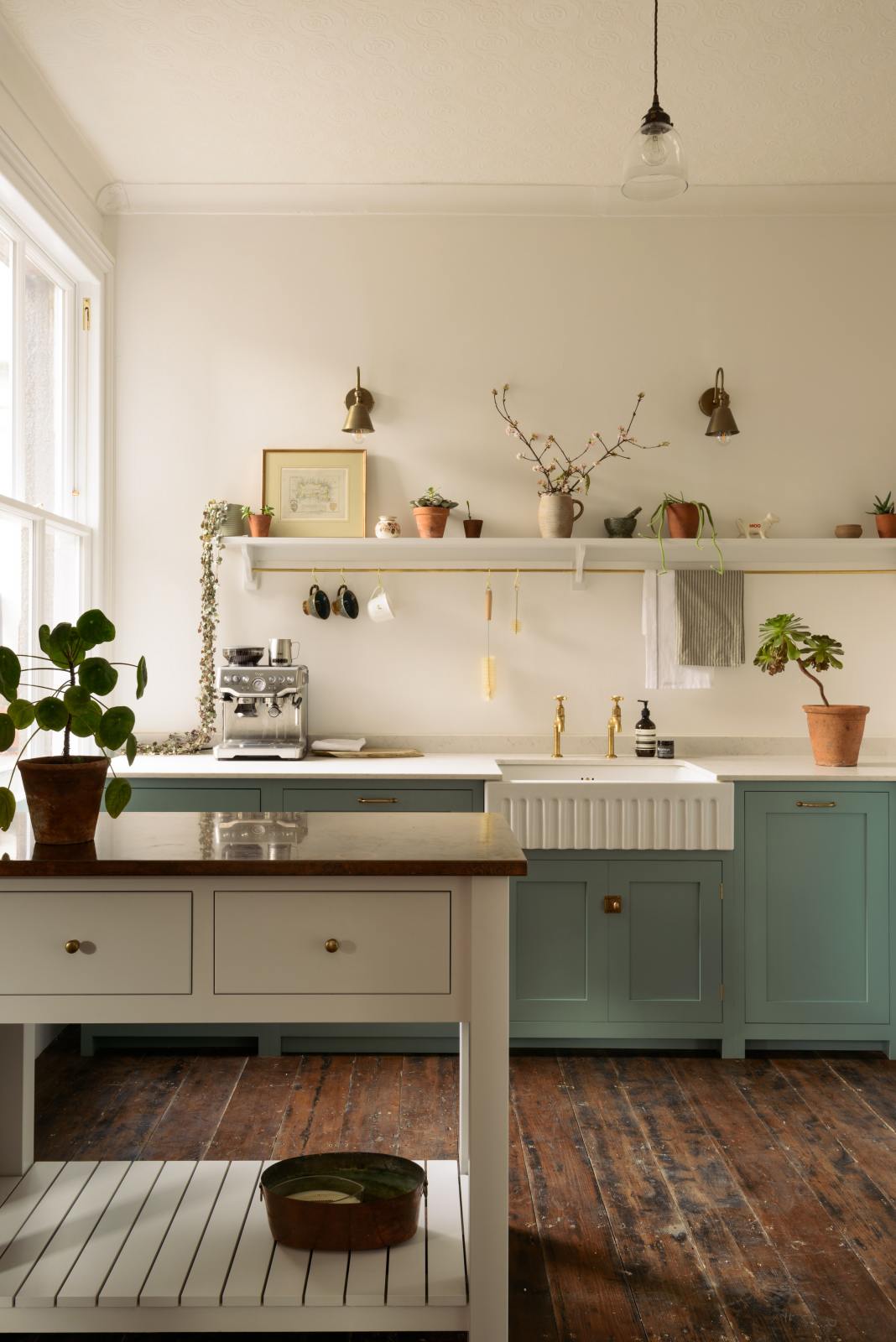 A close look at a freestanding prep table in linen white with a copper worktop in the Trinity Blue shaker kitchen with an L-shaped cooker and sink run. The kitchen features a countertop cupboard with a Carrara marble top, a long shelf with brass rails for hanging cups, cleaning tools, and towels.