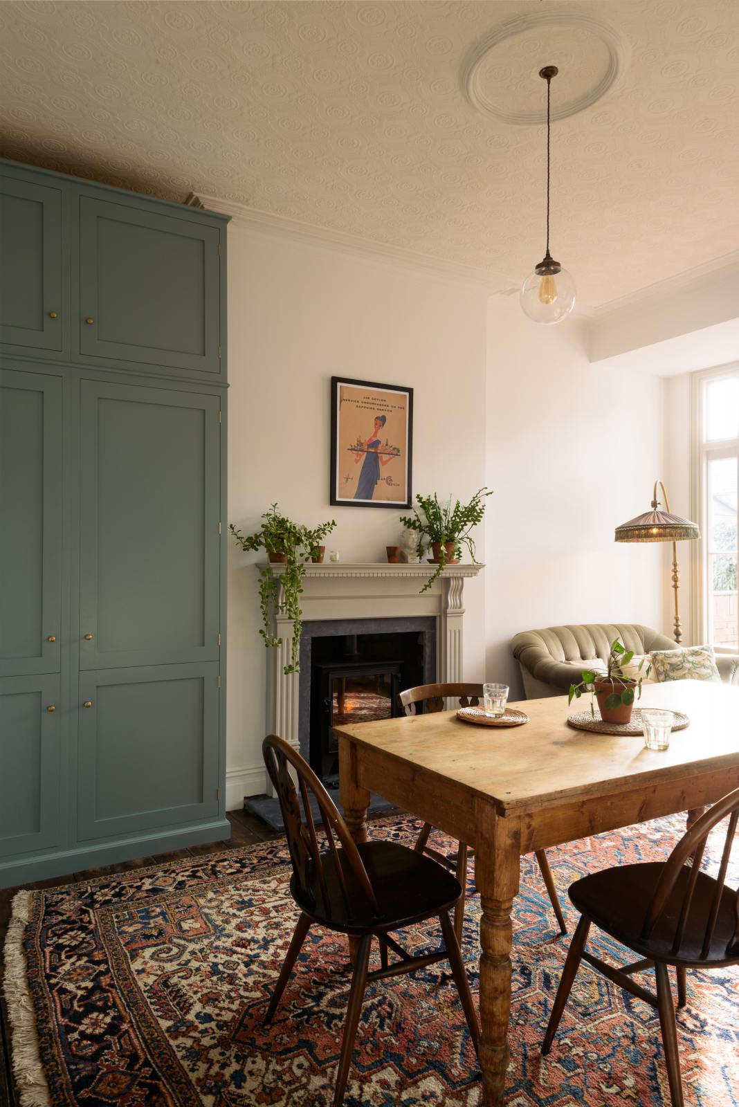 A dining area that is both grand and simple, featuring a vintage table surrounded by mismatched chairs. A Georgian-style fireplace adds character, while a green two-seater sofa and a period lamp sit beside a wide glass window. Blending seamlessly with these found items is a Trinity Blue full-height pantry cupboard.