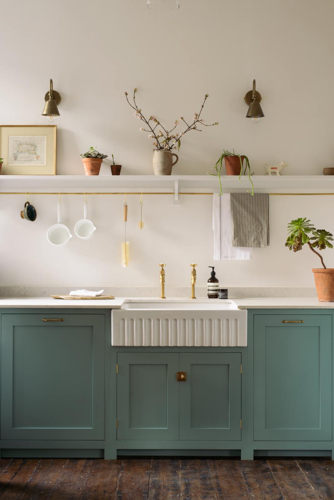 A close-up view of a Trinity Blue shaker kitchen sink features a Carrara marble countertop and a single, fluted Belfast sink with aged brass Mayan taps. Above the sink is a long shelf with brass rails for hanging cups, cleaning tools, and towels.