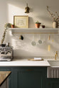 A quaint close-up view of a Trinity Blue shaker kitchen sink features a Carrara marble countertop and a single, fluted Belfast sink with aged brass Mayan taps. Above the sink is a long shelf with brass rails for hanging cups, cleaning tools, and towels.