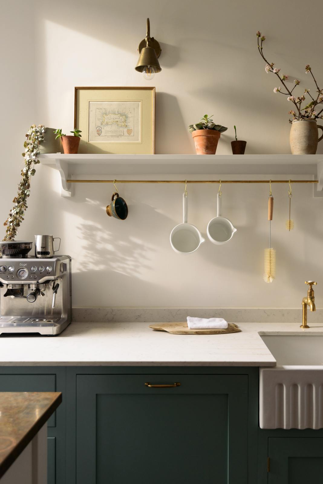 A quaint close-up view of a Trinity Blue shaker kitchen sink features a Carrara marble countertop and a single, fluted Belfast sink with aged brass Mayan taps. Above the sink is a long shelf with brass rails for hanging cups, cleaning tools, and towels.