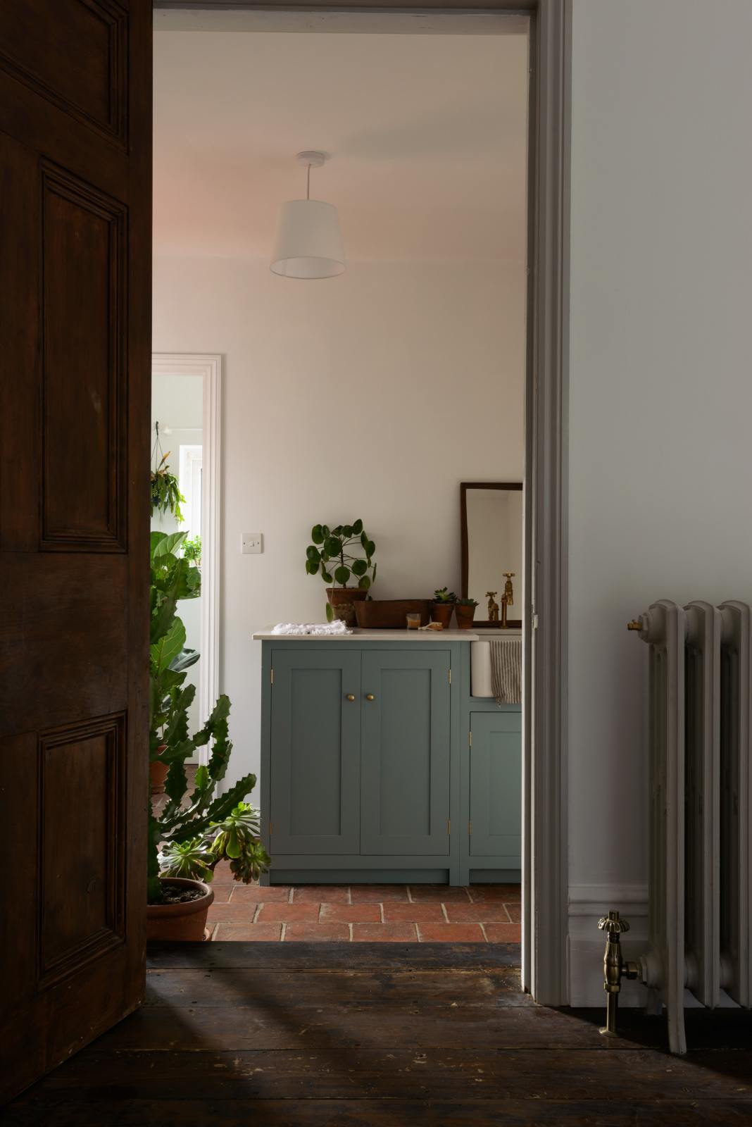 A moody, angled view of a Trinity Blue shaker kitchen sink features a Carrara marble countertop and a single, fluted Belfast sink with aged brass Mayan taps. Above the sink is a long shelf with brass rails for hanging cups, cleaning tools, and towels.