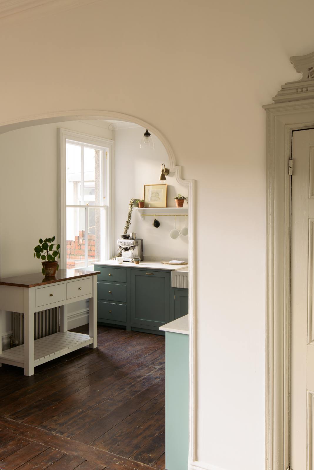 An open door in a well-preserved house with original floorboards reveals a small Trinity Blue scullery room, featuring a Carrara marble countertop, a single Belfast sink with aged brass Mayan taps, and warm terracotta tiles.