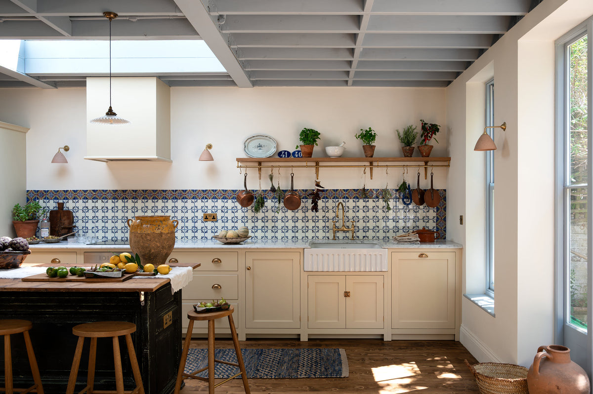 A spacious, Mediterranean-inspired Quaker Cream Shaker kitchen features a long sink and cooker run with a Carrara marble worktop, and a classic oak shelf. Lovingly decorated with a wall of vibrant blue patterned splashback tiles, aged brass hanging rails, Helen’s Stools, green house plants, and fresh produce, it creates a wonderfully social and practical kitchen space.