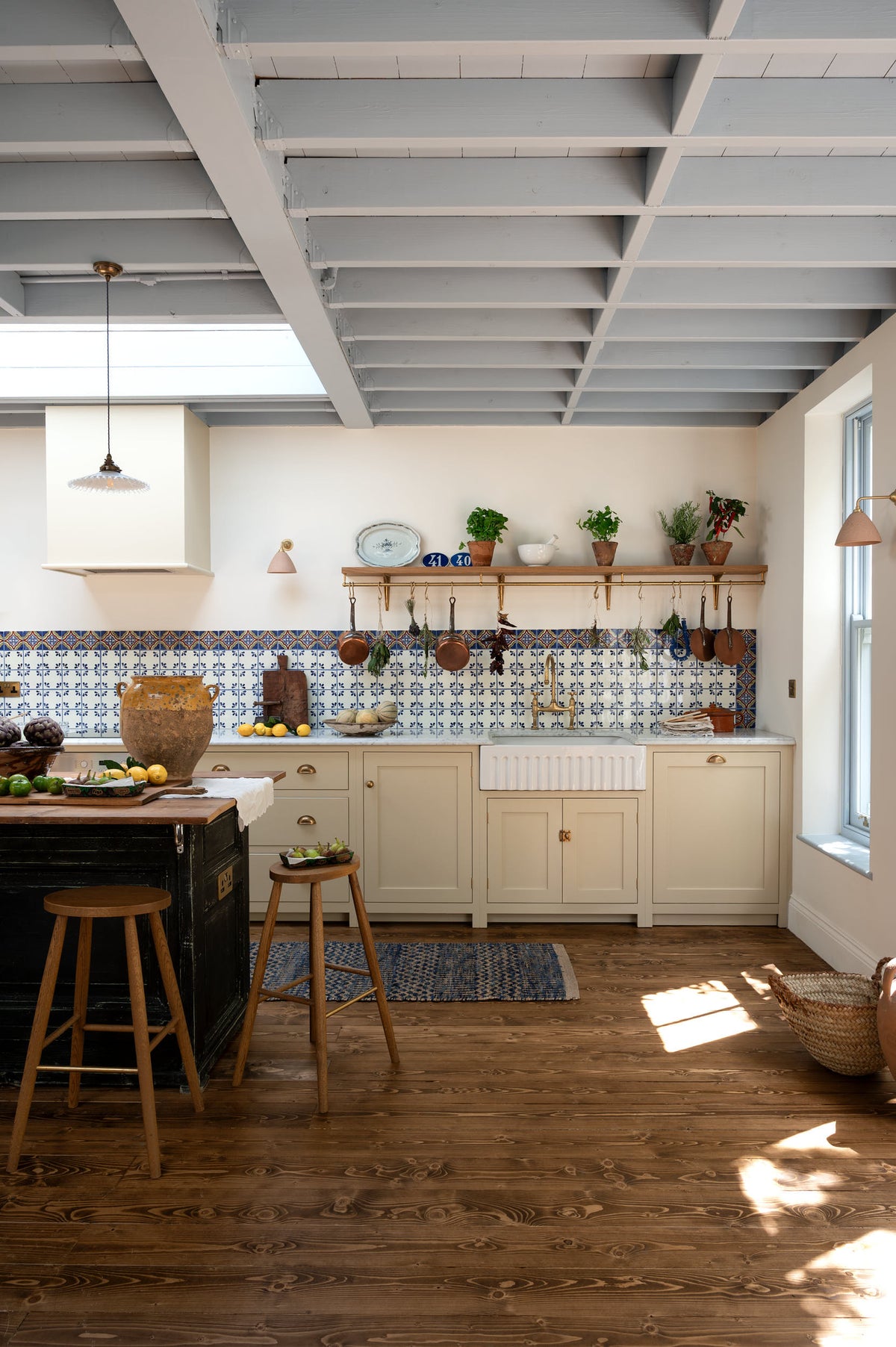 A spacious, Mediterranean-inspired Quaker Cream Shaker kitchen features a long sink and cooker run with a Carrara marble worktop, and a classic oak shelf. Lovingly decorated with a wall of vibrant blue patterned splashback tiles, aged brass hanging rails, Helen’s Stools, green house plants, and fresh produce, it creates a wonderfully social and practical kitchen space.