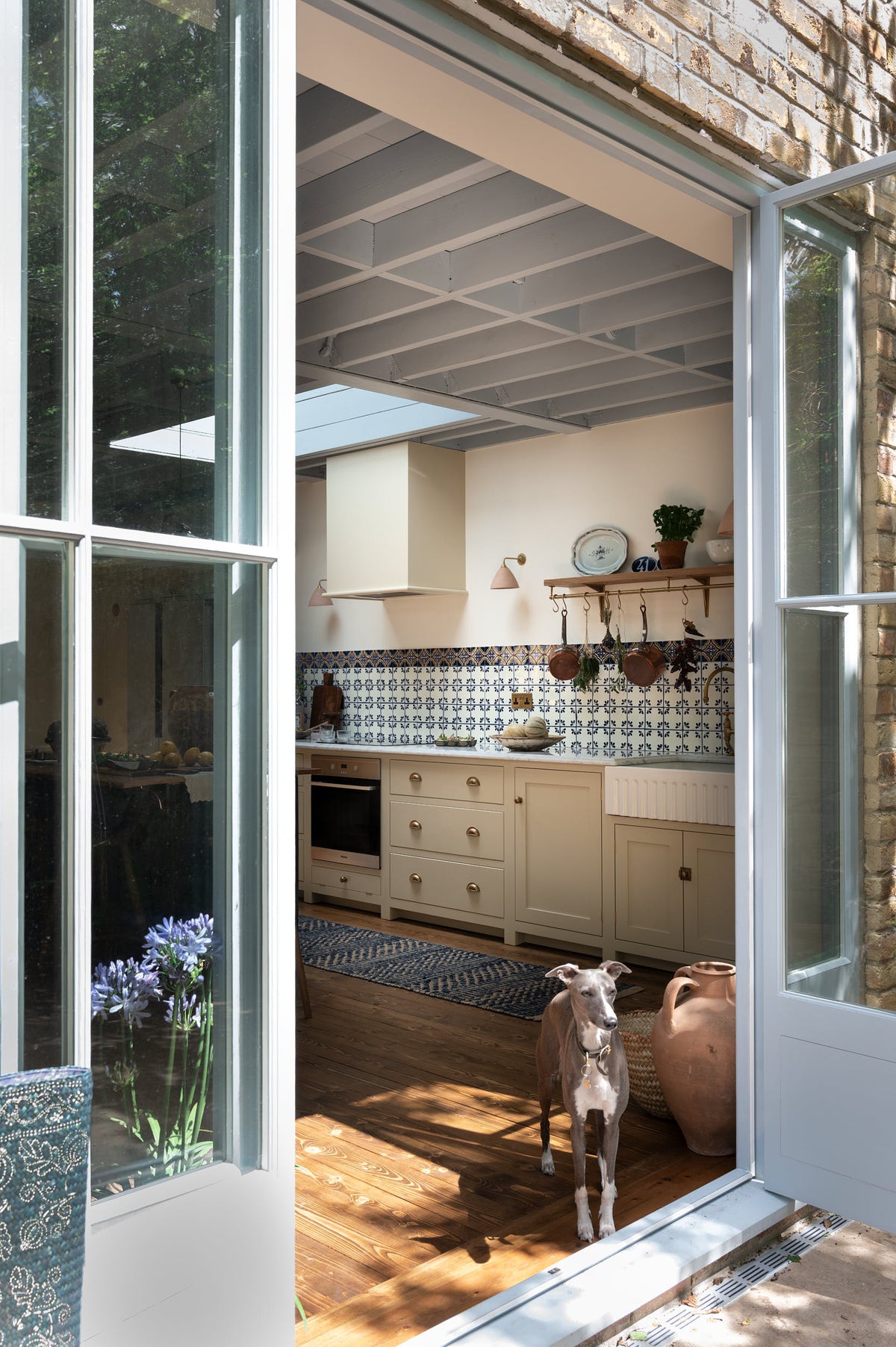 Looking through the powdered blue glass door is a small greyhound puppy, and a spacious, Mediterranean-inspired Quaker Cream Shaker kitchen features a long sink and cooker run with a Carrara marble worktop, and a classic oak shelf. Lovingly decorated with a wall of vibrant blue patterned splashback tiles, aged brass hanging rails, Helen stools, green house plants, and fresh produce, it creates a wonderfully social and practical kitchen space.