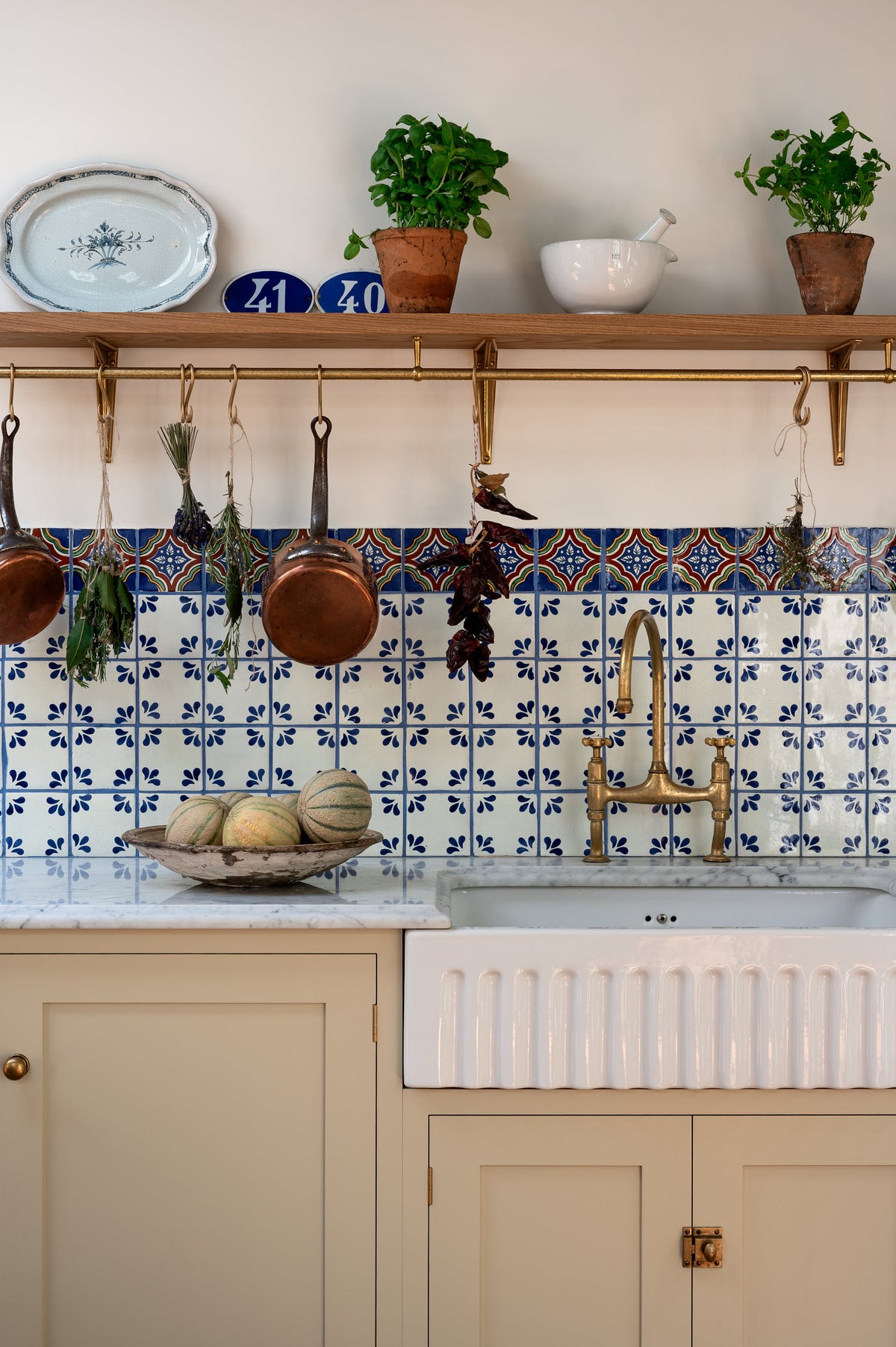 A close-up look at the spacious, Mediterranean-inspired Quaker Cream Shaker kitchen’s sink run with a Carrara marble worktop, and a classic oak shelf. Lovingly decorated with a wall of vibrant blue patterned splashback tiles, aged brass hanging rails, green herbs, an aged brass Ionian tap, and fresh produce, it creates a wonderfully social and practical kitchen space.