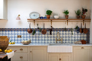A close-up look at the spacious, Mediterranean-inspired Quaker Cream Shaker kitchen’s sink run with a Carrara marble worktop, and a classic oak shelf. Lovingly decorated with a wall of vibrant blue patterned splashback tiles, aged brass hanging rails, green herbs, an aged brass Ionian tap, and fresh produce, it creates a wonderfully social and practical kitchen space.