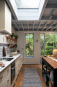 Looking along the galley of the Mediterranean-inspired Quaker Cream Shaker kitchen featuring a long sink and cooker run with a Carrara marble worktop, and a classic oak shelf, it looks through the luscious green garden. Lovingly decorated with a wall of vibrant blue patterned splashback tiles, aged brass hanging rails, green house plants, and fresh produce, it creates a wonderfully social and practical kitchen space.
