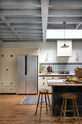 A quiet corner of a spacious, Mediterranean-inspired Quaker Cream Shaker kitchen features a long sink and cooker run with a Carrara marble worktop, a pantry cupboard, and a classic oak shelf. Lovingly decorated with a wall of vibrant blue patterned splashback tiles, aged brass hanging rails, Helen stools, green house plants, and fresh produce, it creates a wonderfully social and practical kitchen space.