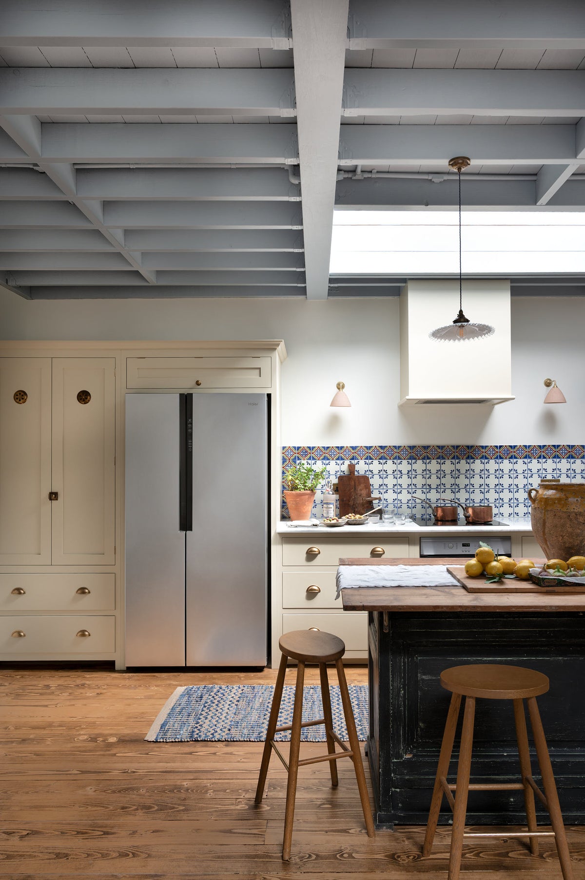 A quiet corner of a spacious, Mediterranean-inspired Quaker Cream Shaker kitchen features a long sink and cooker run with a Carrara marble worktop, a pantry cupboard, and a classic oak shelf. Lovingly decorated with a wall of vibrant blue patterned splashback tiles, aged brass hanging rails, Helen stools, green house plants, and fresh produce, it creates a wonderfully social and practical kitchen space.