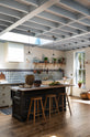 An oblique view of a spacious, Mediterranean-inspired Quaker Cream Shaker kitchen features a long sink and cooker run with a Carrara marble worktop, and a classic oak shelf. Lovingly decorated with a wall of vibrant blue patterned splashback tiles, aged brass hanging rails, Helen stools, green house plants, and fresh produce, it creates a wonderfully social and practical kitchen space.