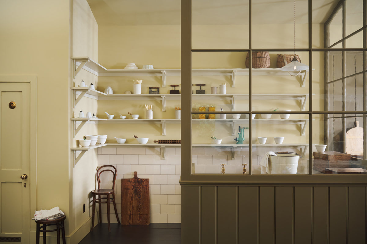 A simple Quaker cream and Folgate green Shaker scullery features architectural glazed partitions, bespoke shelving, and a sink run with a Silestone worktop, finished a cream subway tiles splashback. Lovingly adorned with artisan ceramics, wooden chopping boards, and antique kitchen accessories and furniture, it gives a sombre and practical space.