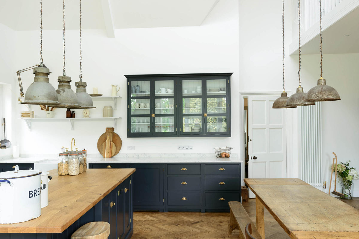 A tall pantry-blue shaker kitchen features a sink run with a Silestone worktop, paired with a large island topped with solid oak and glazed cupboards. A vintage oak dining table with wooden benches and mismatched chairs adds character, while rusty metal pendant lights in the centre bring an industrial edge.