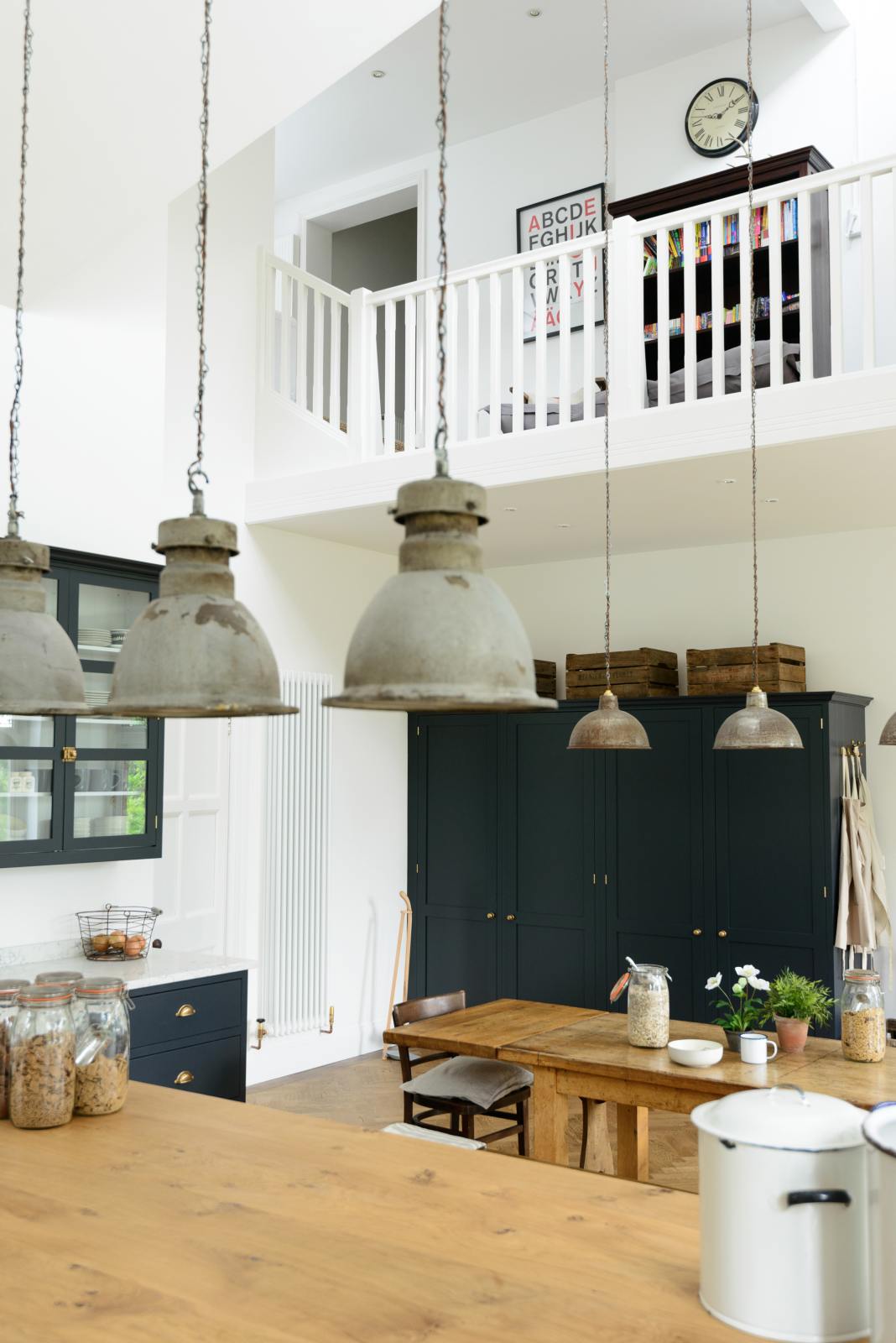 High ceilings with a balcony overlook the large pantry-blue shaker kitchen, featuring a sink run with a Silestone worktop, glazed cupboards, a solid oak-topped island, and two full-height pantry cupboards. A vintage oak dining table with wooden benches and mismatched chairs adds character, while rusty metal pendant lights in the centre bring an industrial edge.