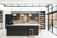 A corner of a bright and spacious pantry-blue Shaker kitchen under a wall-to-ceiling skylight features a long cooker run with wall cabinets, a kitchen island with a sink, and a concrete worktop. Adorned with glass pendant lights, collected artworks, a bowl of shallots, and everyday kitchen items, it creates a warm and social space.