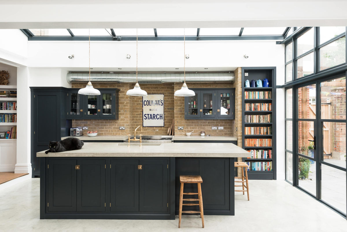A corner of a bright and spacious pantry-blue Shaker kitchen under a wall-to-ceiling skylight features a long cooker run with wall cabinets, a kitchen island with a sink, and a concrete worktop. Adorned with glass pendant lights, collected artworks, a bowl of shallots, and everyday kitchen items, it creates a warm and social space.